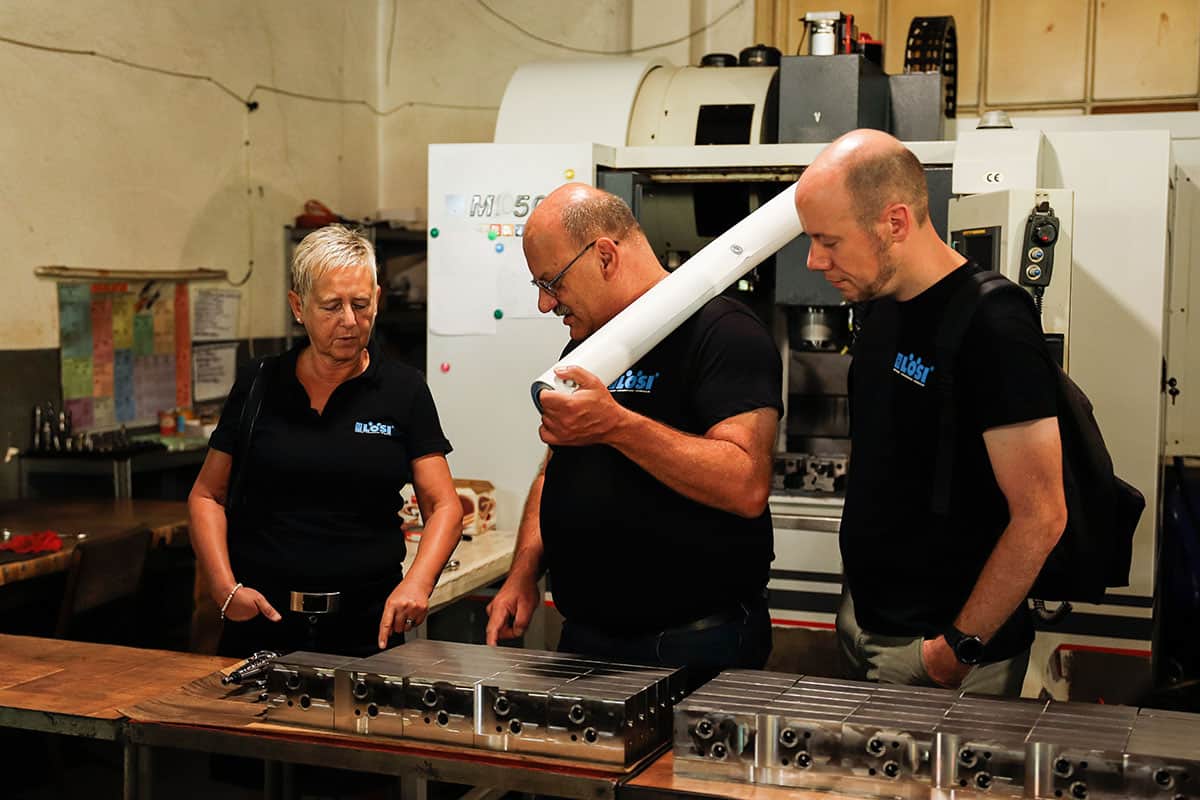 Three LöSi employees wearing black work polo shirts inspect milled parts on a work table in a workshop hall. A CNC milling machine can be seen in the background.